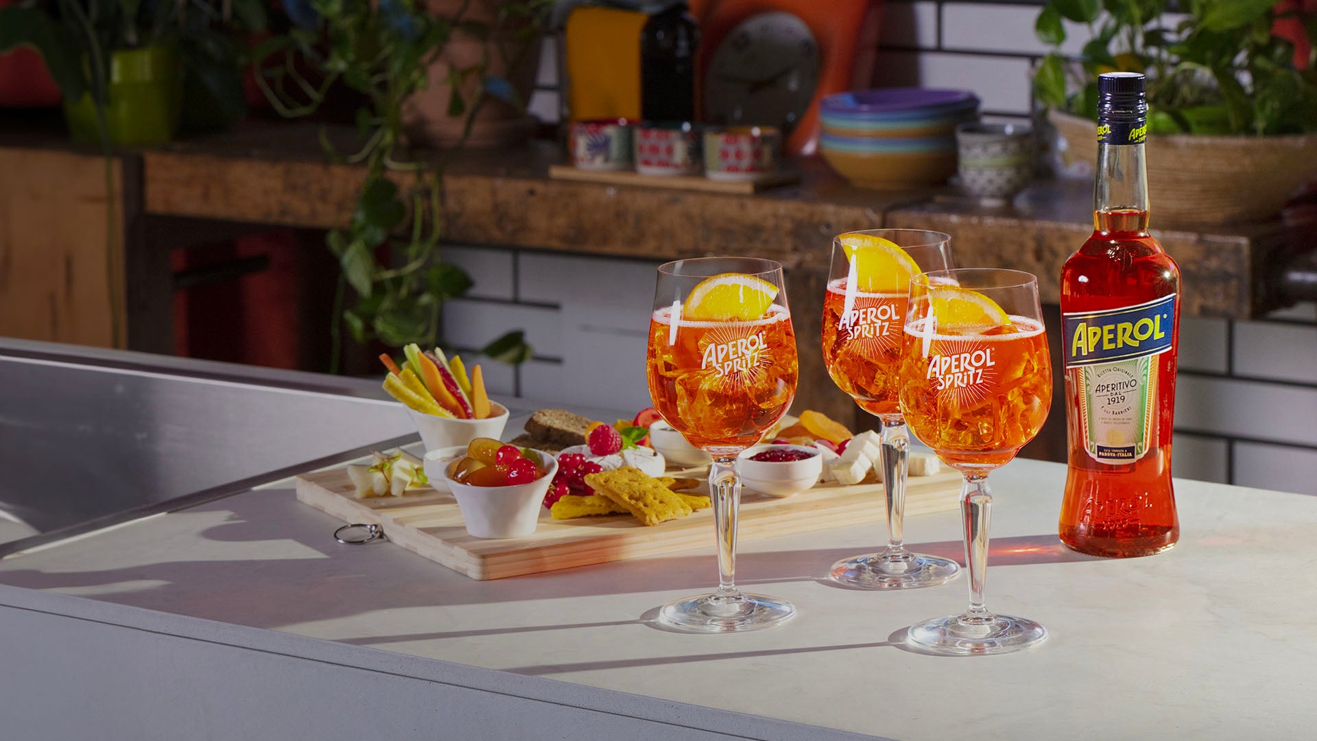 Three Aperol Spritz cocktails garnished with orange slices are placed on a kitchen counter next to a full bottle of Aperol and a wooden platter filled with colorful appetizers, creating a vibrant aperitivo setting.